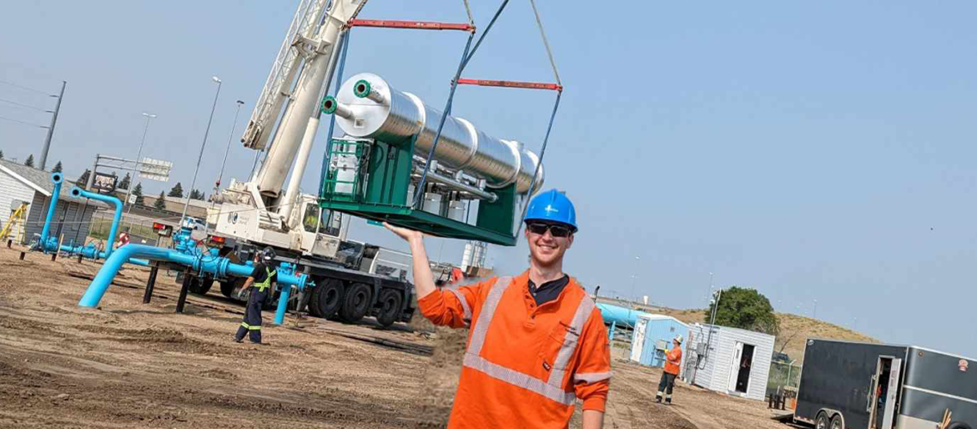 A man wearing an orange workshirt and blue hard hat holds up his arm, to make it appear he is lifting a large silver line heater on a crane in the background.