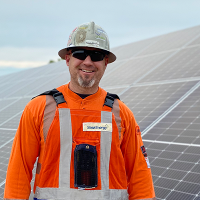 A man in a grey hardhat and orange long-sleeved safety shirt stands in front of solar panels, with blue sky and a tree beyond. 